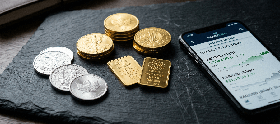Gold and silver coins and small gold bars on a dark slate surface with a smartphone displaying live gold and silver prices